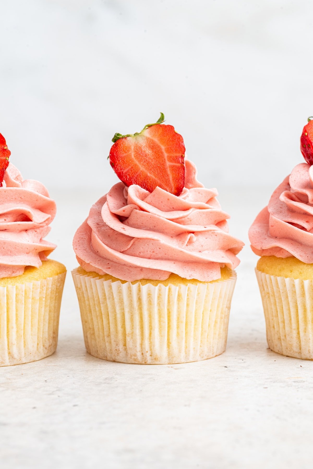 A vanilla cupcake with a pink strawberry buttercream frosting and a fresh strawberry slice on top.