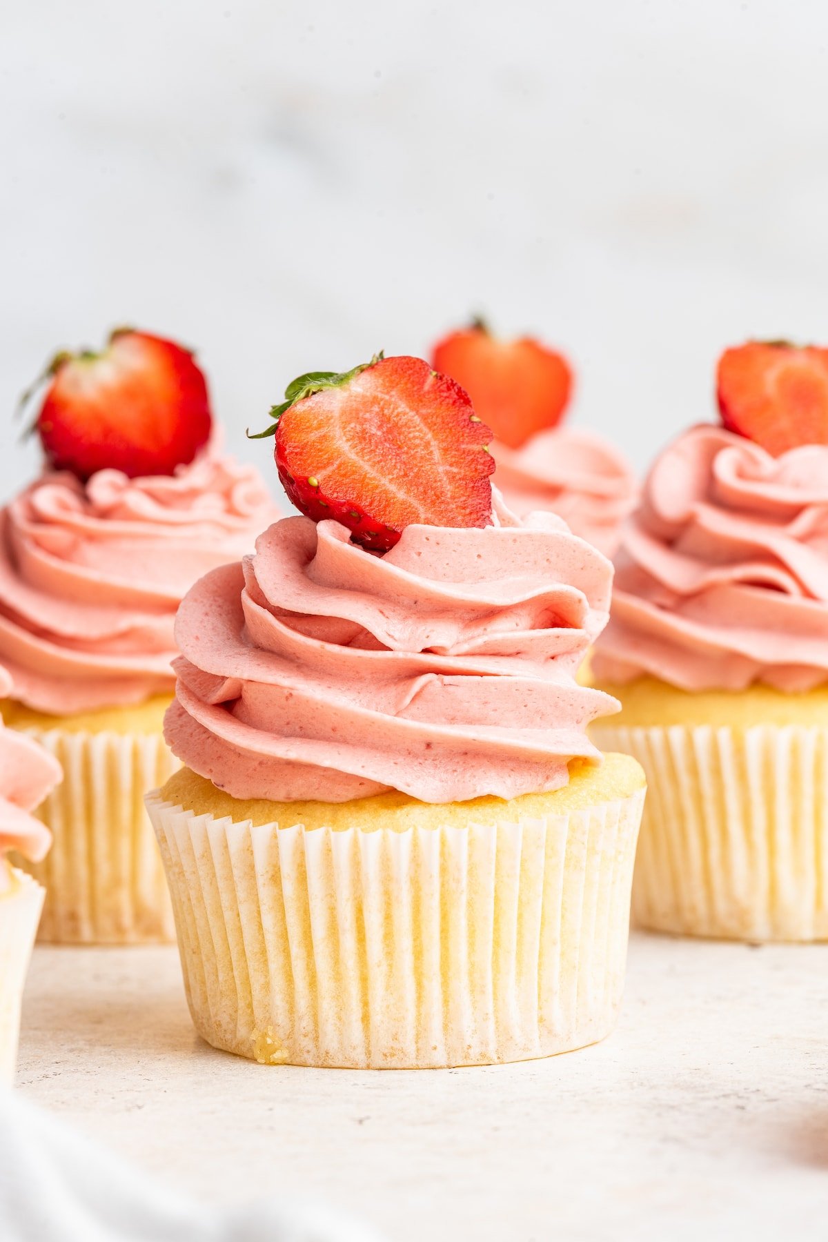 A cupcake standing center, with others in the background. The cupcakes are topped with a pink frosting and fresh strawberry slices on top.