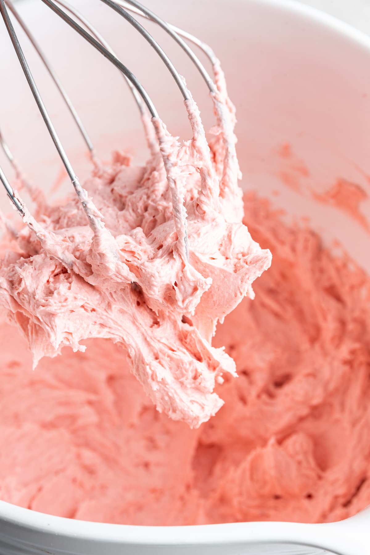 A whisk leaves a white bowl full of pink strawberry buttercream frosting.