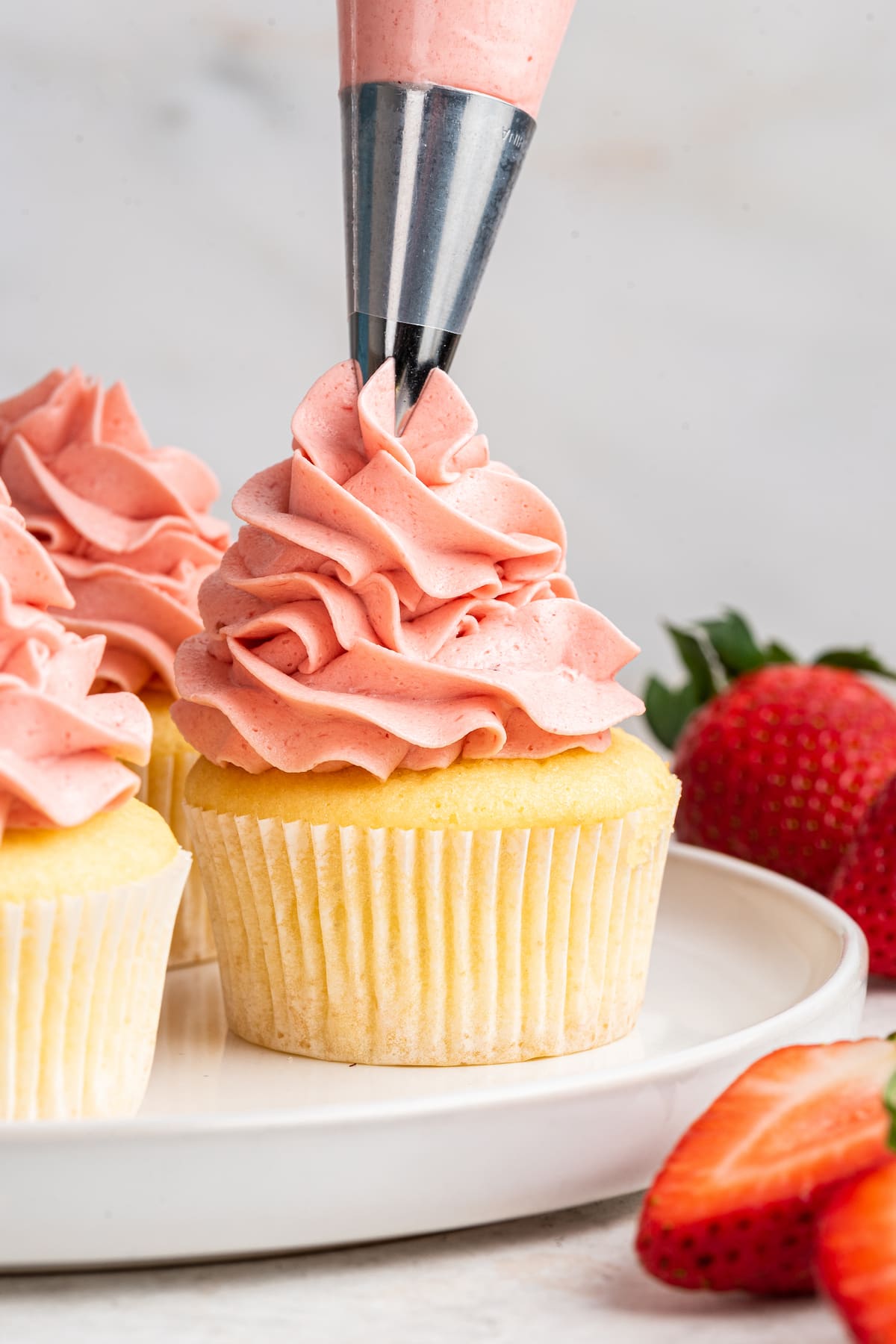 A vanilla cupcake on a white plate being topped off with strawberry buttercream frosting from a pastry bag.