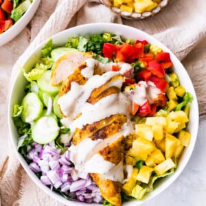 A white bowl containing cooked sliced chicken on top of a bed of lettuce, red onion, cucumber, red bell pepper, pineapple, and a creamy dijon mustard dressing.