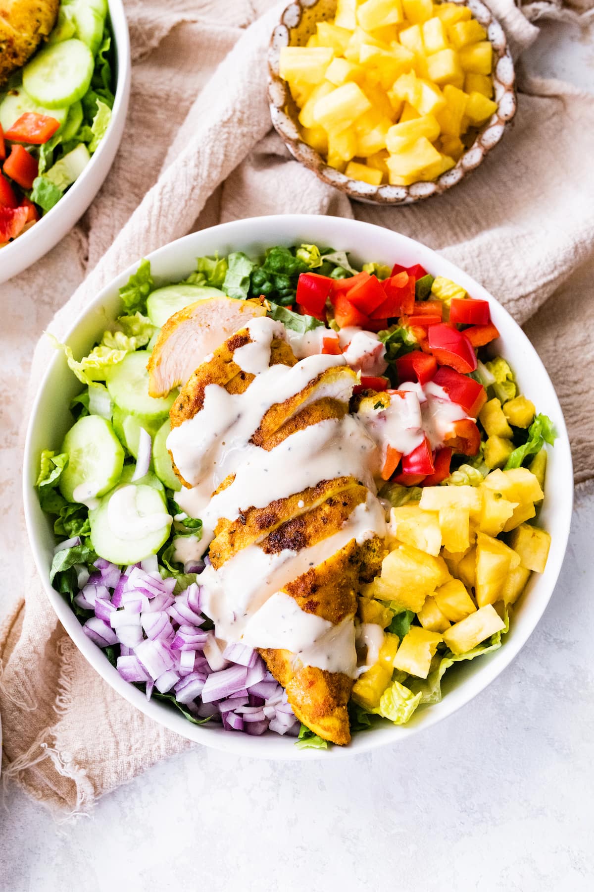 A white bowl containing cooked sliced chicken on top of a bed of lettuce, red onion, cucumber, red bell pepper, pineapple, and a creamy dijon mustard dressing.