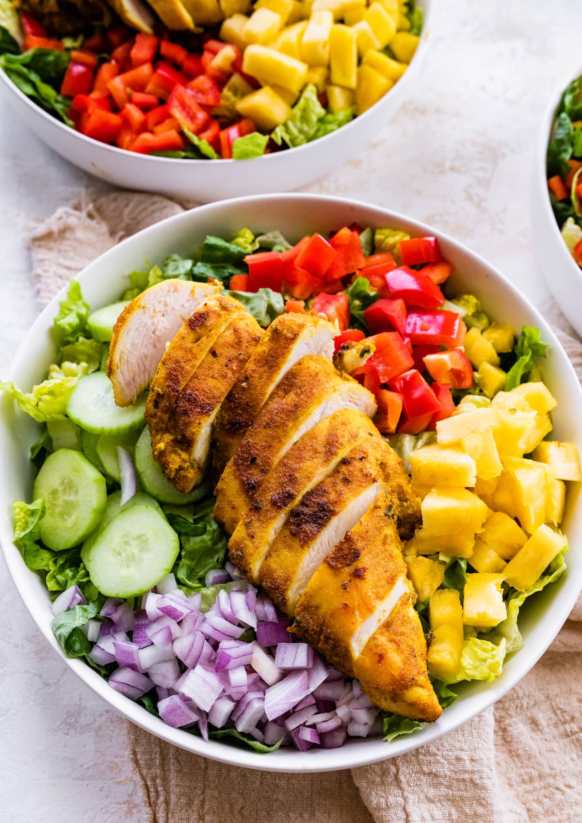 A white bowl containing cooked sliced chicken on top of a bed of lettuce, red onion, cucumber, red bell pepper, and pineapple.