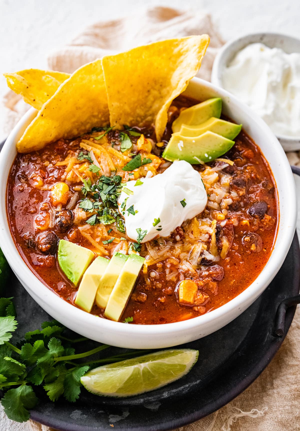 A white bowl full of turkey chili, topped sour cream, shredded cheese, avocado slices, fresh cilantro, and tortilla chips.