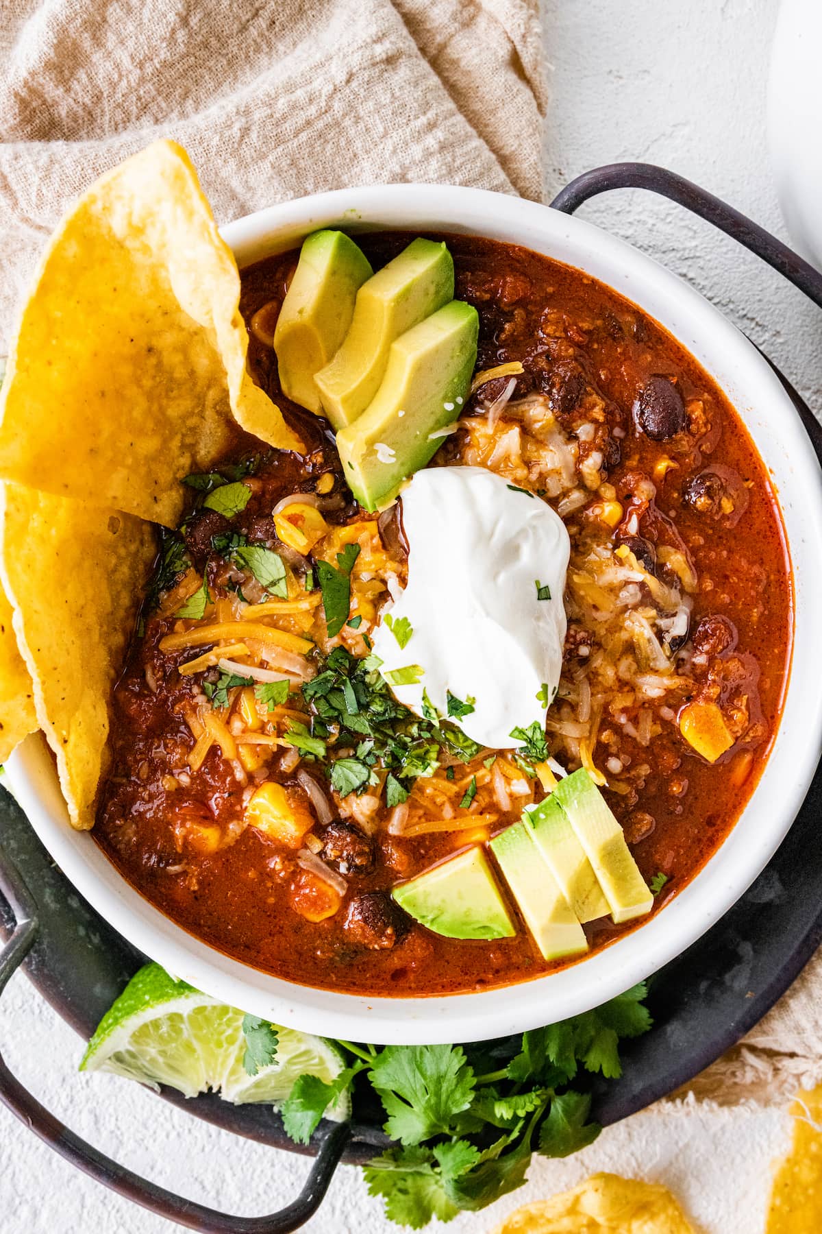 A white bowl full of turkey chili, topped sour cream, shredded cheese, avocado slices, fresh cilantro, and tortilla chips.