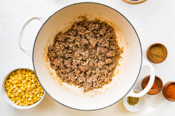 Cooked ground turkey in a large white dutch oven. There's a small white bowl full of corn on one side, and four small cups with spices on the other side.