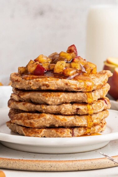 Five apple pancakes stacked on one another on a white plate with diced apples and maple syrup on top.