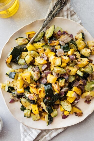 Roasted zucchini and squash on a white plate with a metal spoon.