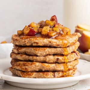 Five apple pancakes stacked on one another on a white plate with diced apples and maple syrup on top.