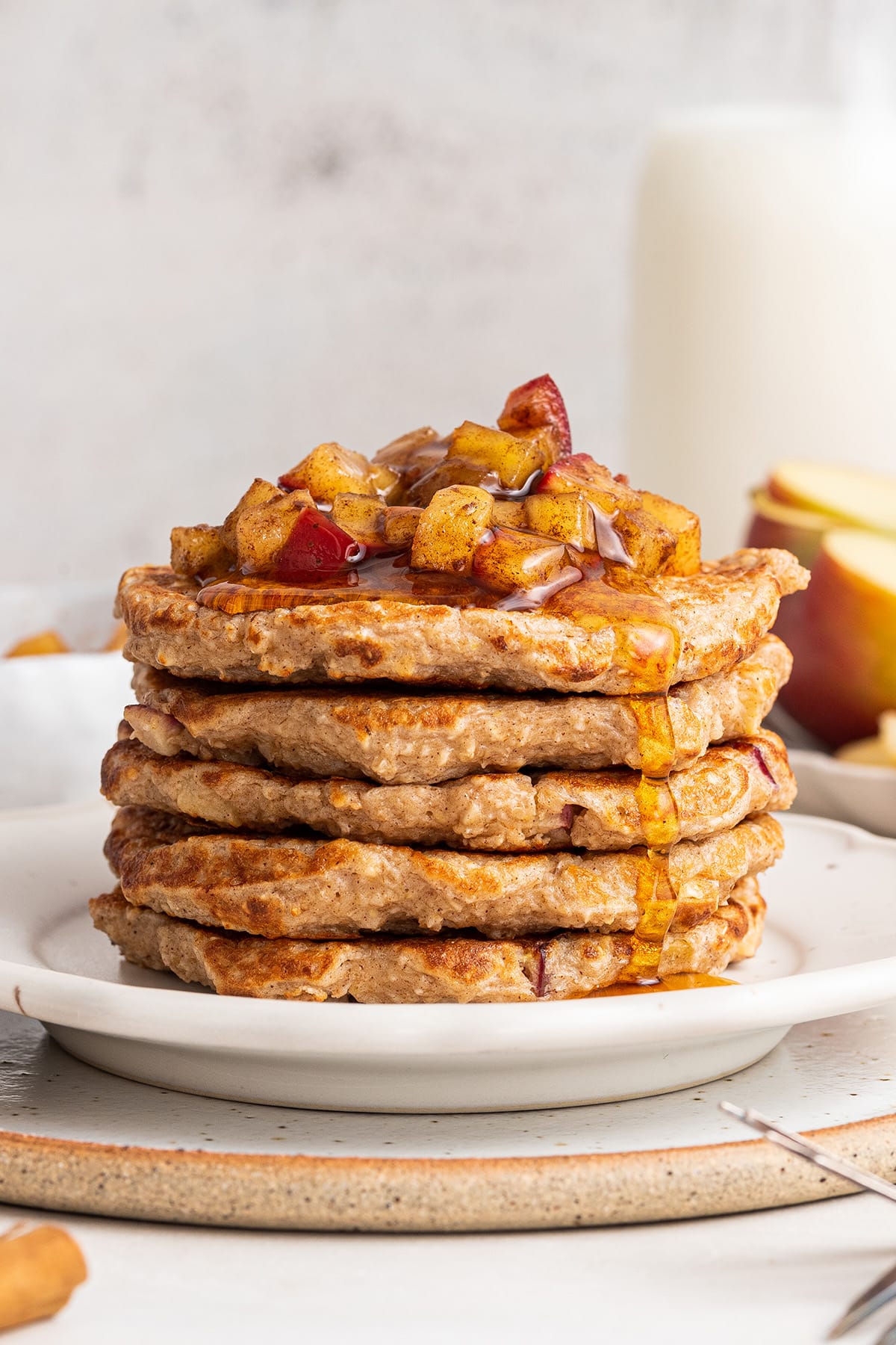 Five apple pancakes stacked on one another on a white plate with diced apples and maple syrup on top.