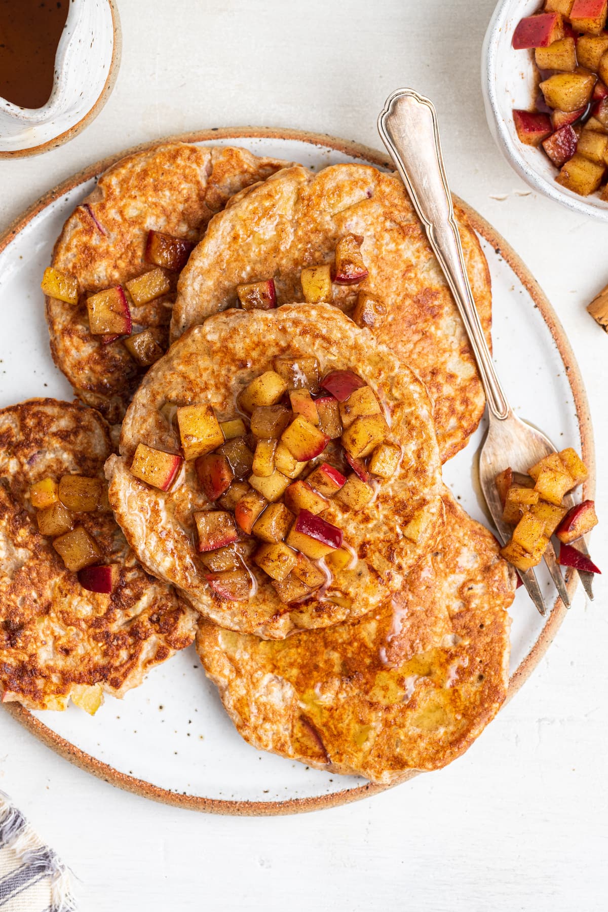 Five apple pancakes on a white plate with a metal fork and diced apples with maple syrup on top.