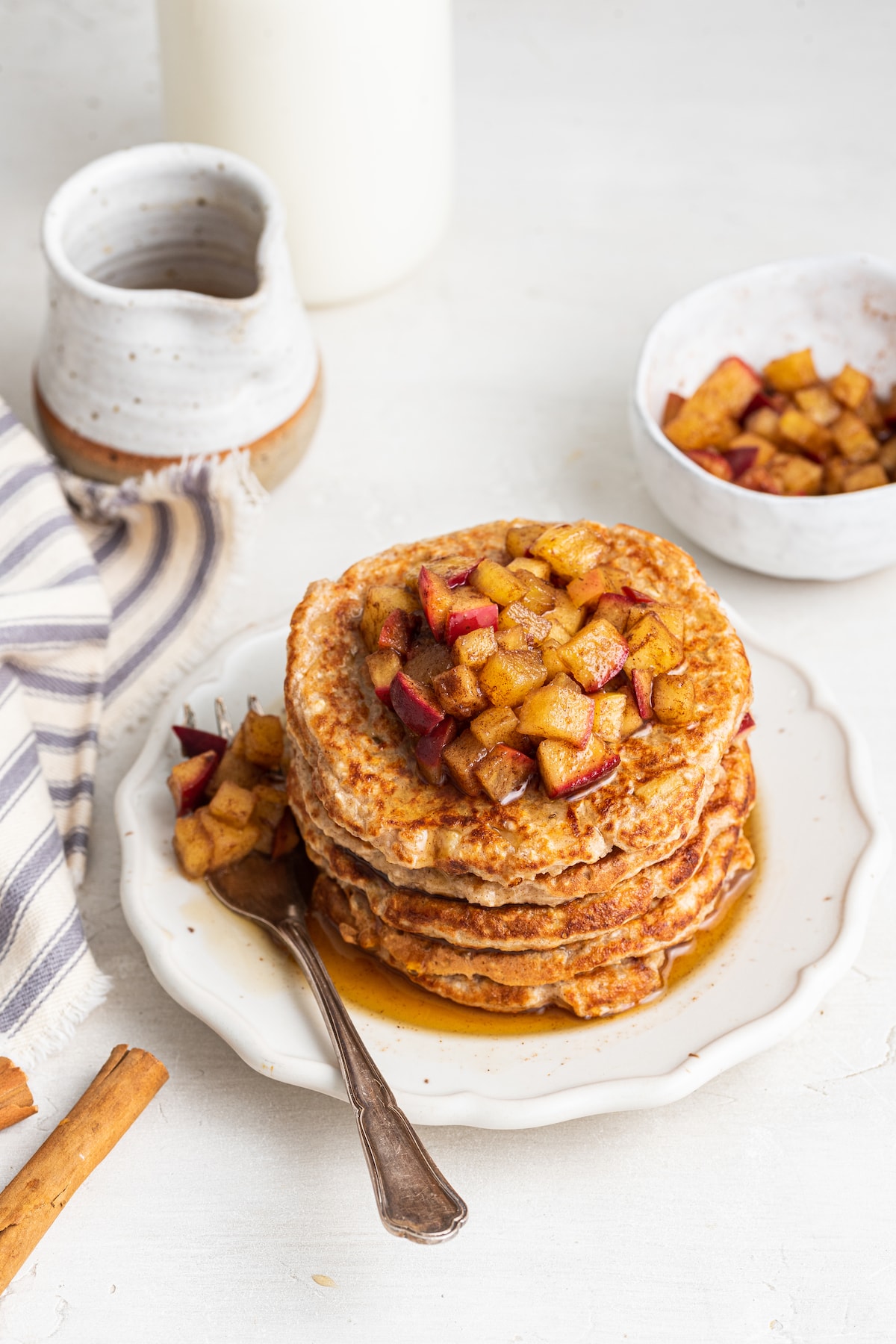 Five apple pancakes stacked on one another on a white plate with a metal fork, with diced apples and maple syrup on top.