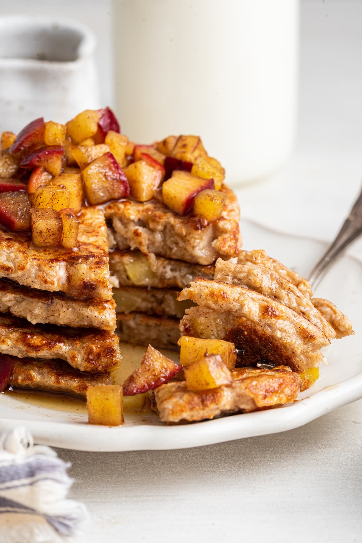 Stacked apple pancakes on a white plate with a bite-size piece on a fork in front of the pancakes.