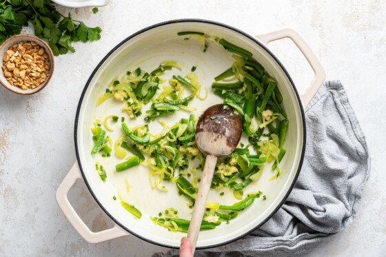 Green pepper, yellow pepper, and onion in a large white pot with a woman's hand holding a wooden spoon.