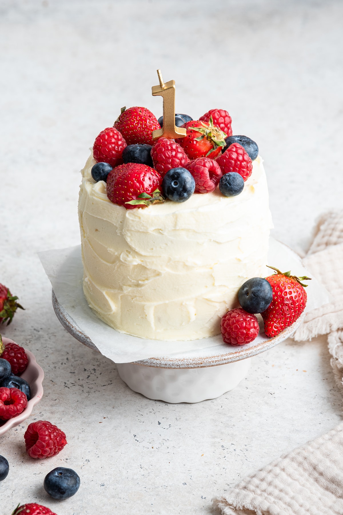 A frosted smash cake with a one candle topped with fresh berries sitting on a cake stand.