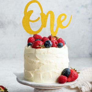 A frosted smash cake with a one sign topped with fresh berries sitting on a cake stand.