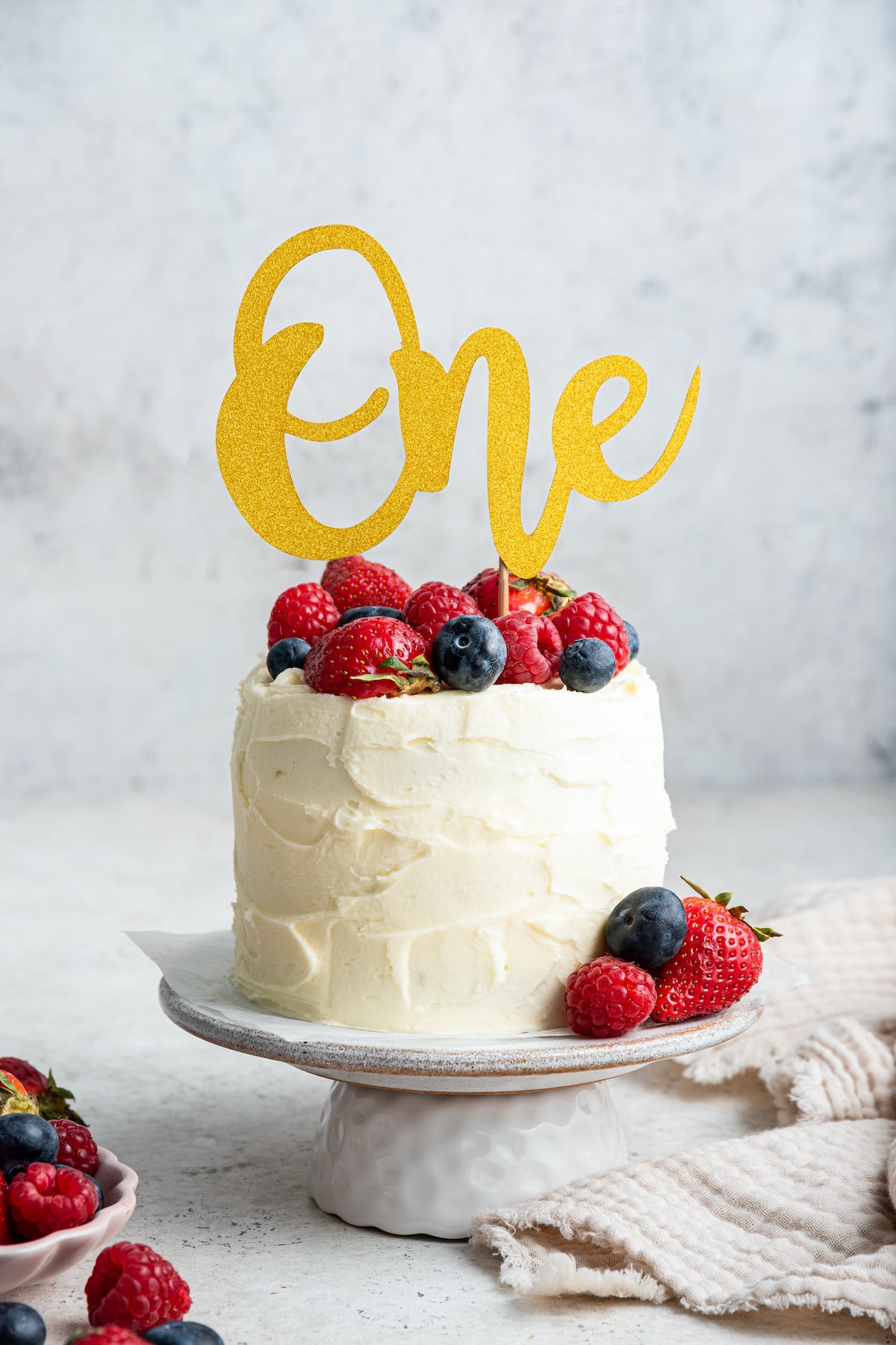 A frosted smash cake with a one sign topped with fresh berries sitting on a cake stand.