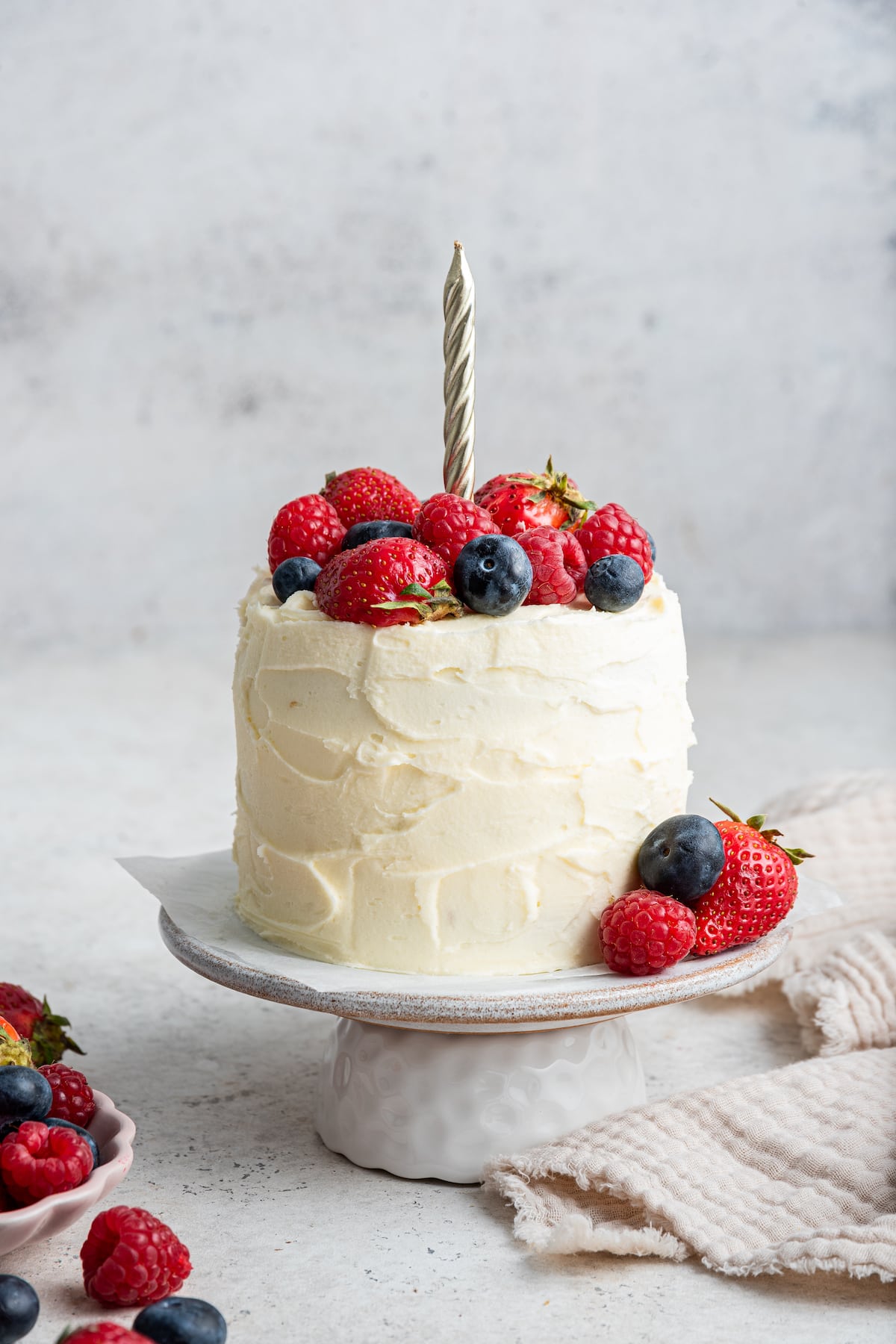 A frosted smash cake with a candle topped with fresh berries sitting on a cake stand.