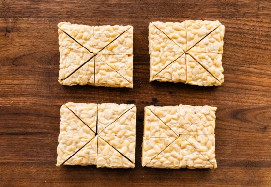 Raw tempeh on a wooden cutting board, cut into triangles.