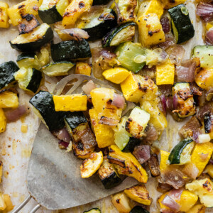 Roasted zucchini, squash, and red onion on a baking tray with a metal spatula.