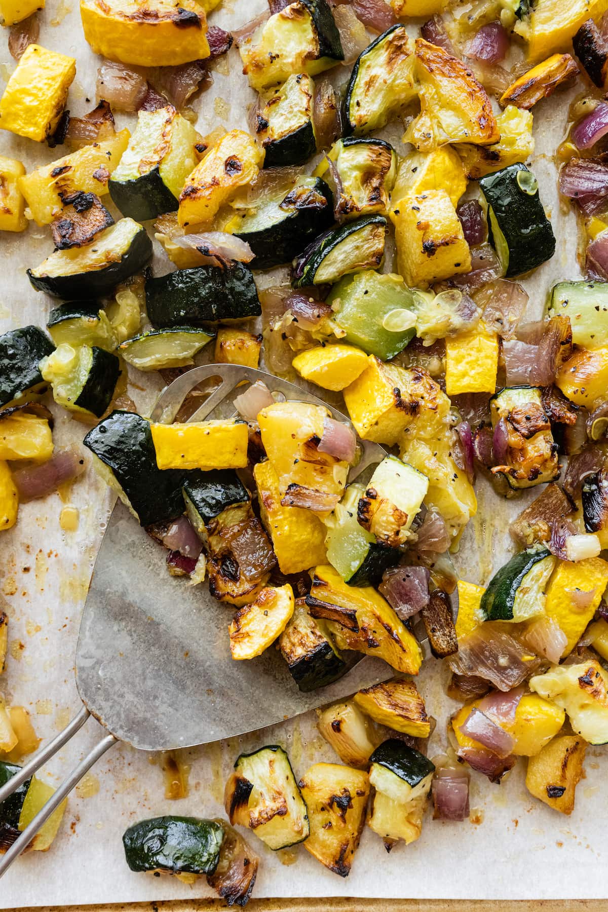 Roasted zucchini, squash, and red onion on a baking tray with a metal spatula.