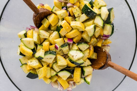 A glass bowl with zucchini, squash, red onion, spices, and two wooden spoons.