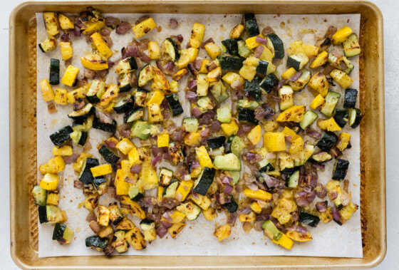 Roasted zucchini, squash, garlic, and red onion on a baking tray.