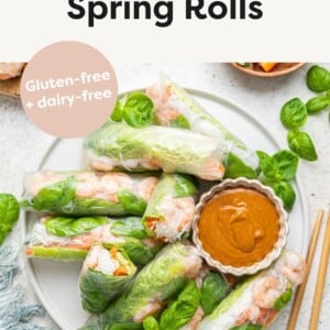 A large white plate containing multiple shrimp spring rolls, some cut in half, and a peanut sauce in a small white bowl. Wooden chopsticks are near the plate, and fresh basil leaves are used as a garnish.