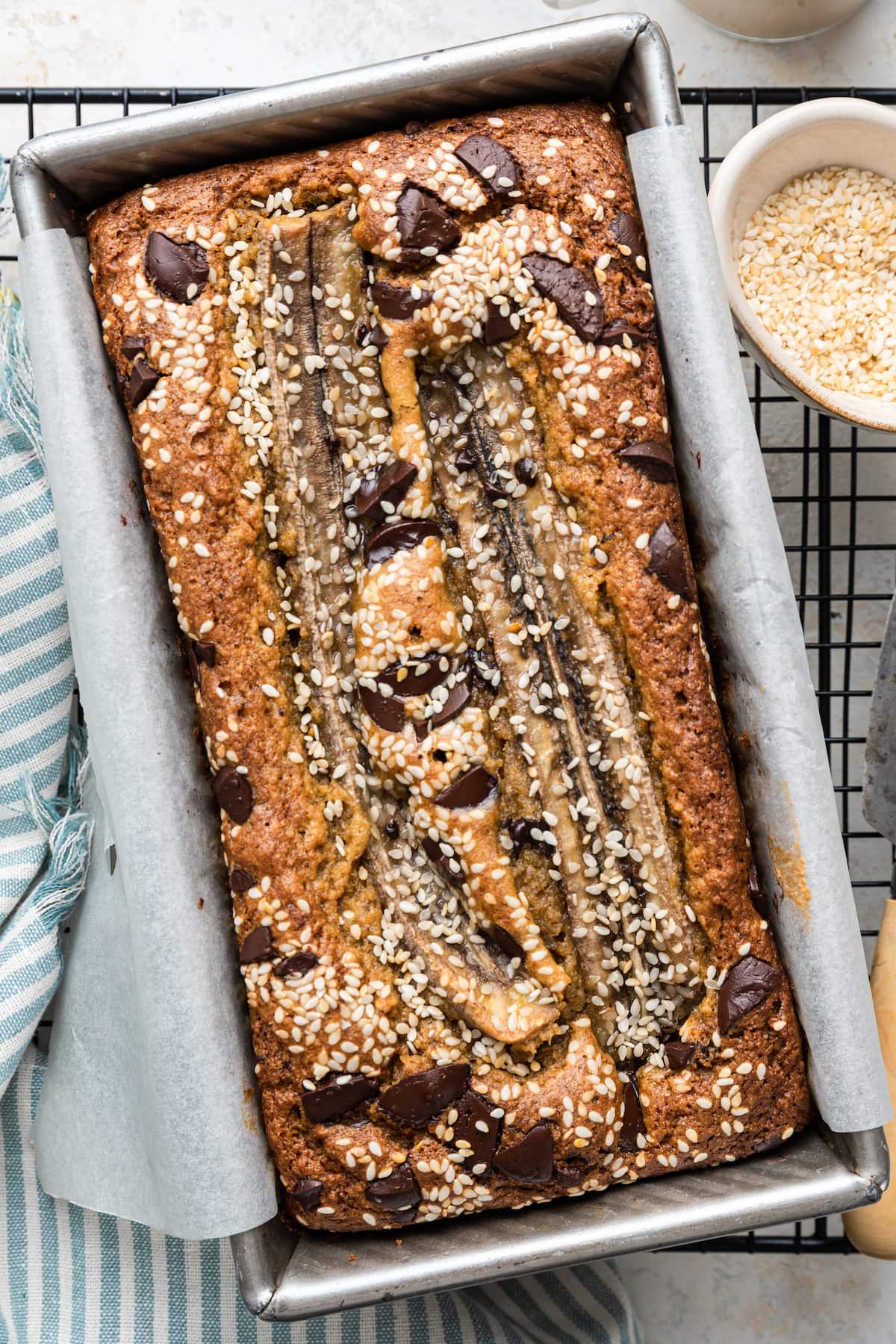 A top view of tahini banana bread inside a bread pan on a wire rack after being baked in the oven. The bread is topped with sesame seeds and chocolate chips.