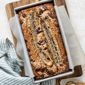 A top view of tahini banana bread inside a bread pan on a wooden cutting board after being baked in the oven. The bread is topped with sesame seeds and chocolate chips.