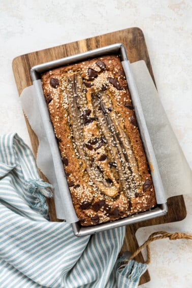 A top view of tahini banana bread inside a bread pan on a wooden cutting board after being baked in the oven. The bread is topped with sesame seeds and chocolate chips.