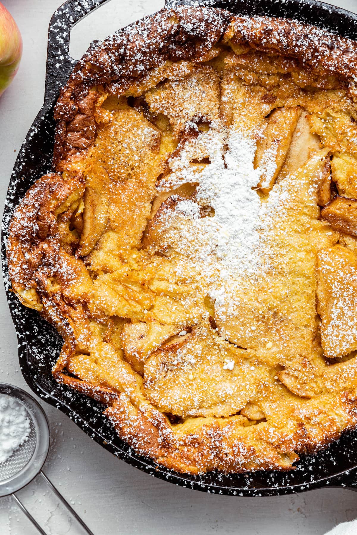 A close up of the cooked apple dutch baby in a cast iron pan sprinkled with powdered sugar.