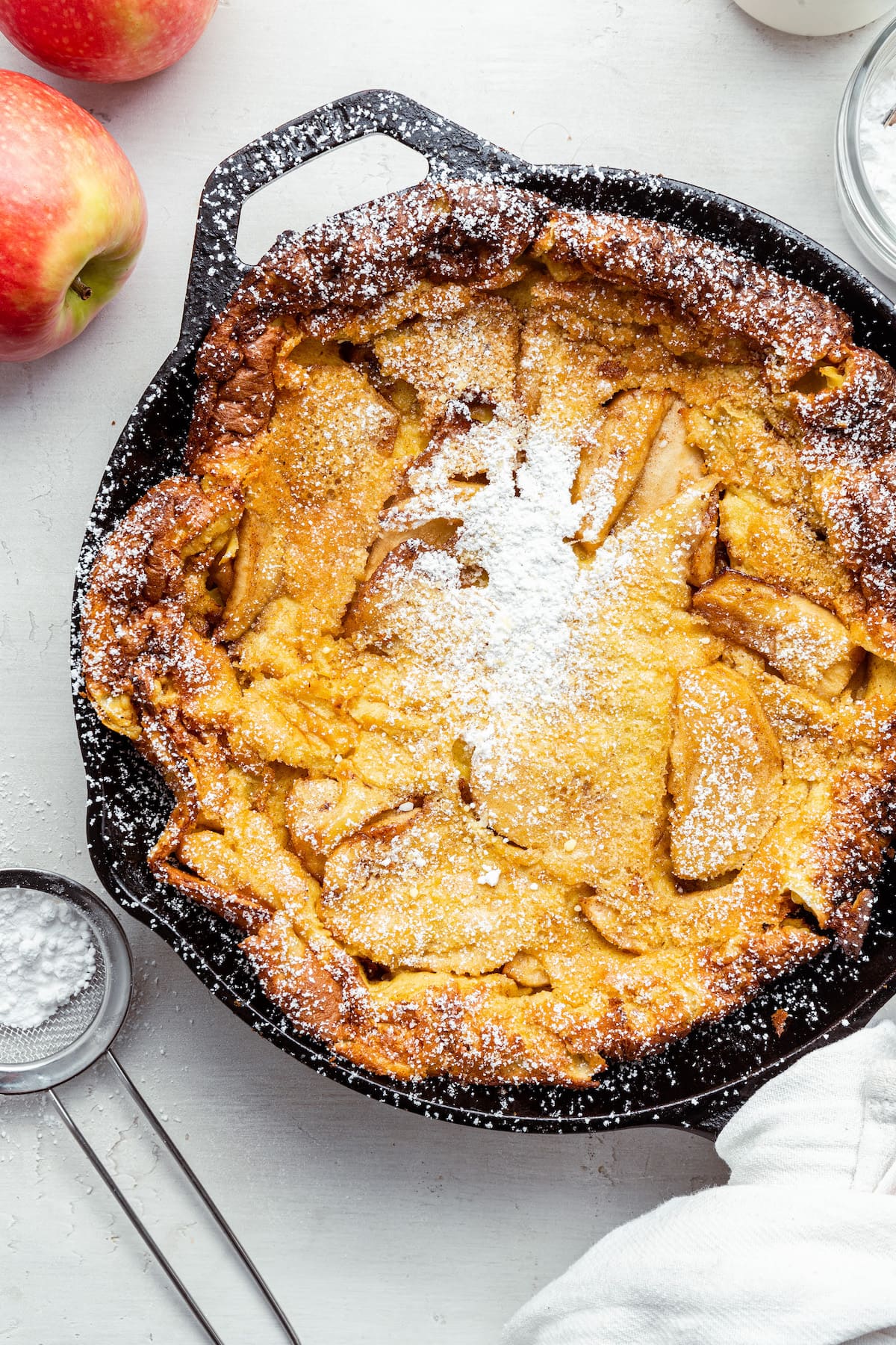 A cooked apple dutch baby in a cast iron with powdered sugar sprinkled on top.