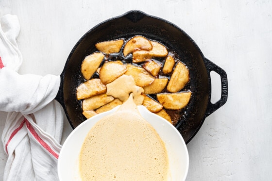 The wet ingredients being poured into the cast iron pan with the cinnamon apples to cook the apple dutch baby.