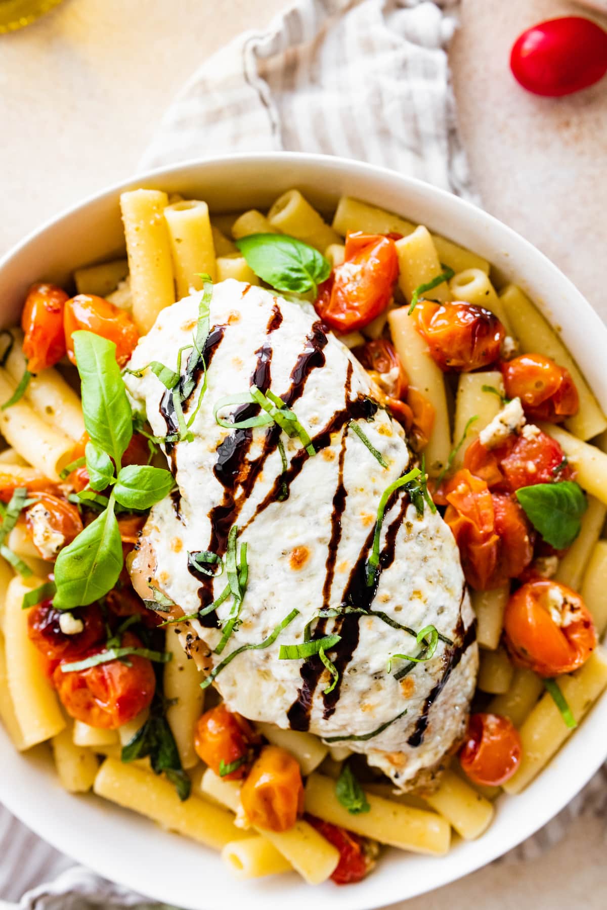 A baked caprese chicken breast with melted mozzarella and a balsamic glaze on top of a bowl of pasta.