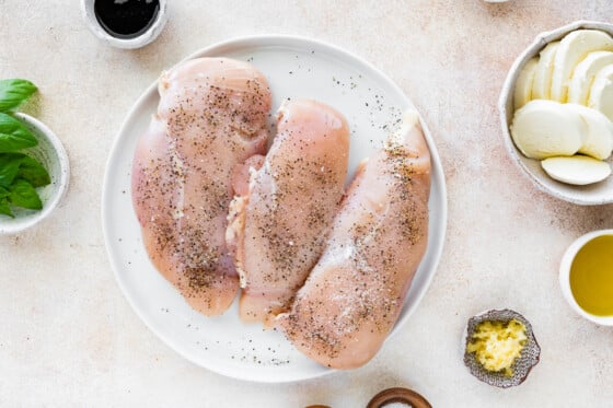 Three raw chicken breasts on a large plate seasoned with salt and pepper.