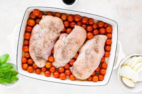 Three raw chicken breasts in a large baking dish over a bed of cherry tomatoes and oil.