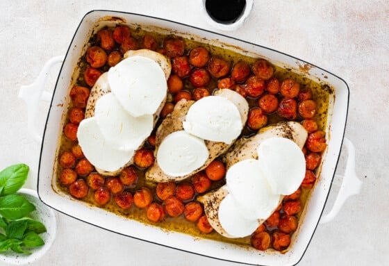 Three chicken breasts on a bed of cherry tomatoes in a baking dish after being baked and the chicken breasts are topped with fresh mozzarella.