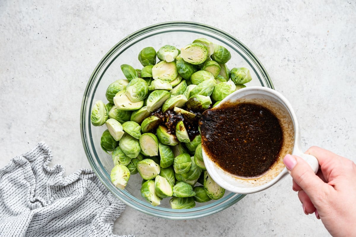 A woman's hand pouring a balsamic mixture over halved Brussels sprouts in a large glass bowl