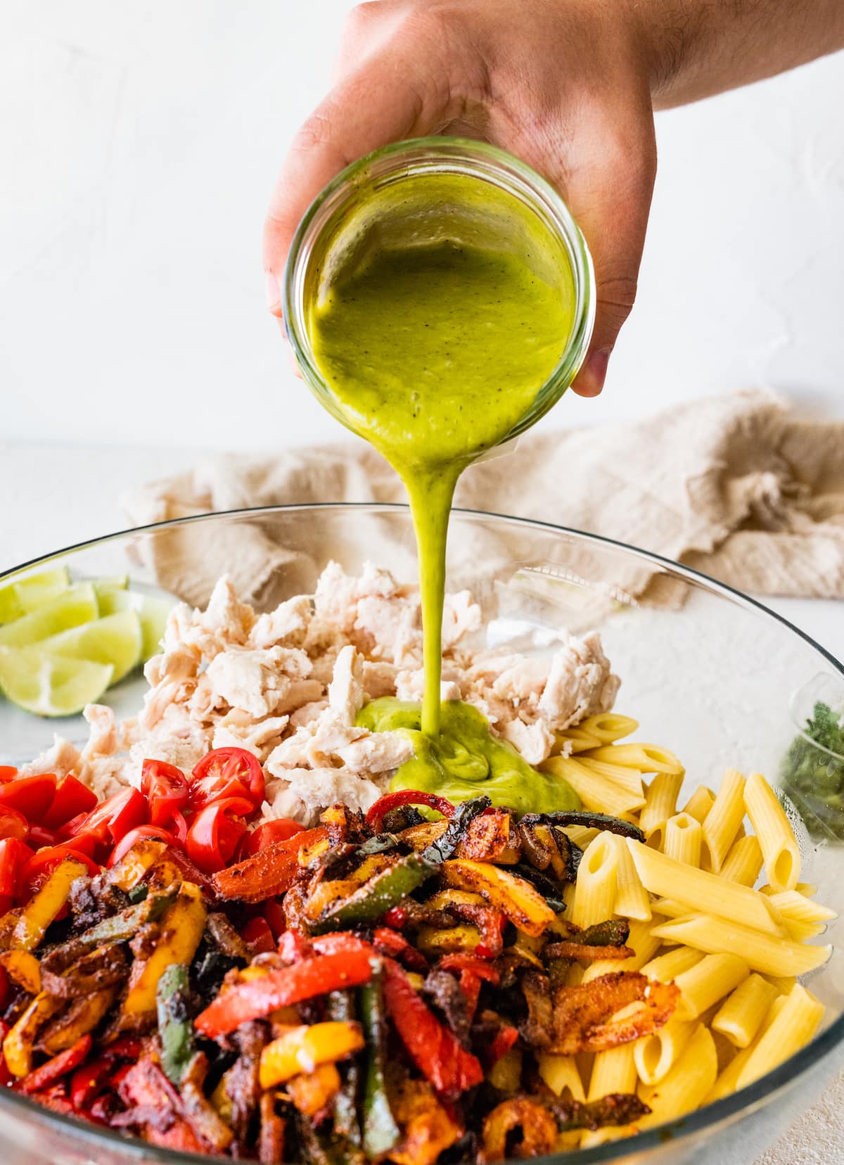 A woman's hand pouring a cilantro lime dressing into a glass bowl of a chicken fajita pasta salad.