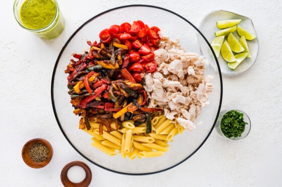 Chicken fajita pasta salad ingredients in a large glass bowl. Noticeable ingredients are; chicken, cherry tomatoes, penne pasta, and cooked bell peppers.