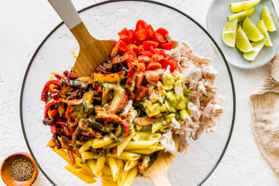 Chicken fajita pasta salad that is about to be mixed with two wooden serving spoons in a large glass bowl.