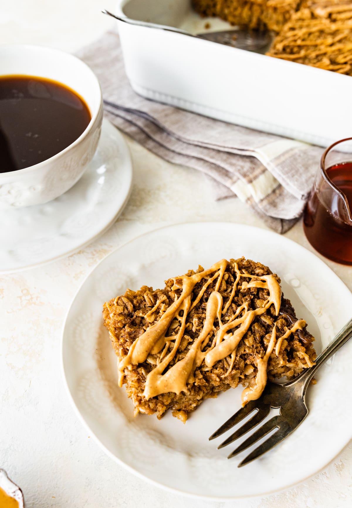A slice of coffee baked oatmeal on a small plate with a metal fork near a cup of coffee.