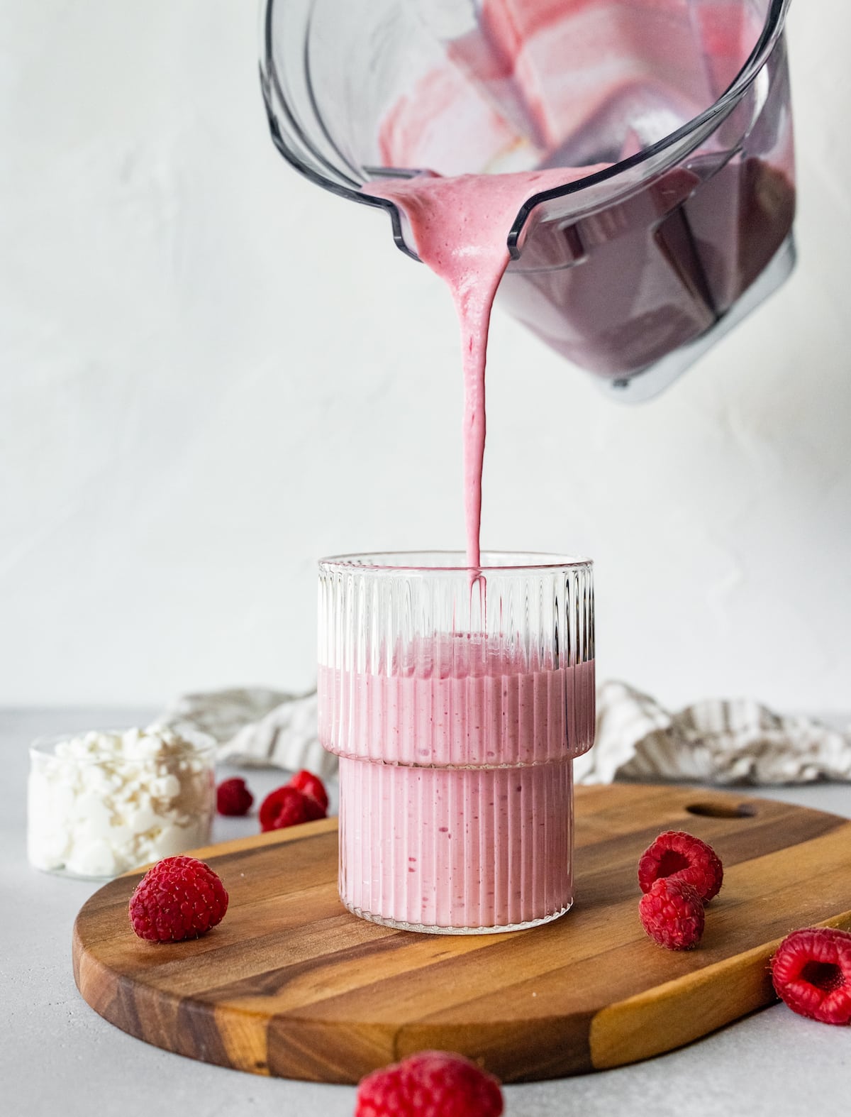 A cottage cheese smoothie being poured into a glass from the blender. Glass is sitting on a wooden cutting board with fresh raspberries scattered around.