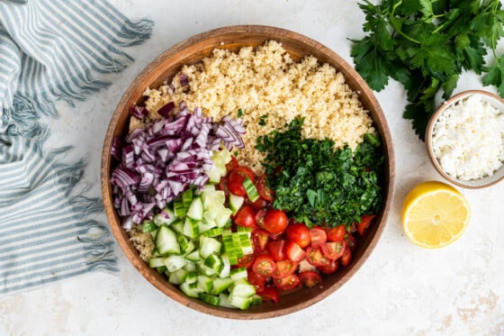 All ingredients for the couscous salad in a large wooden bowl. Noticeable ingredients include couscous, red onion, parsley, cherry tomatoes, and cucumber.