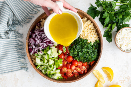A woman's hand pouring a lemon dressing over a couscous salad in a large wooden bowl.