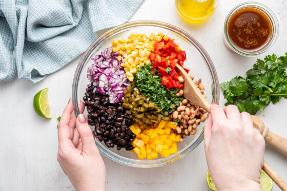 A woman's hand using a wooden spoon to mix the ingredients of the cowboy caviar in a large glass bowl.