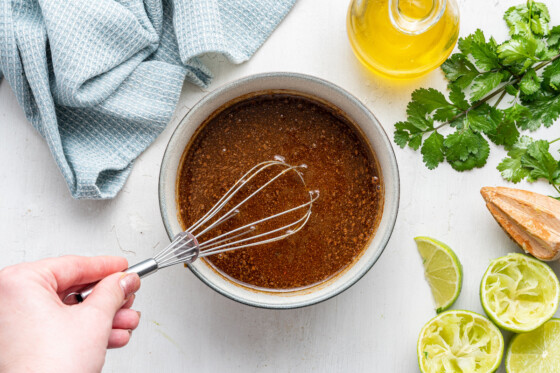 A woman's hand using a metal whisk to whisk a bowl of homemade dressing.