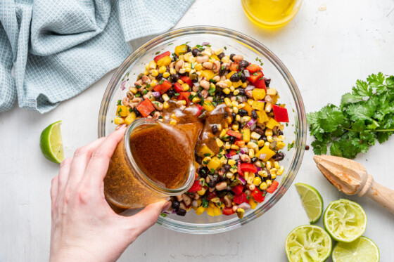 A woman's hand pouring a dressing into a large glass bowl of cowboy caviar.