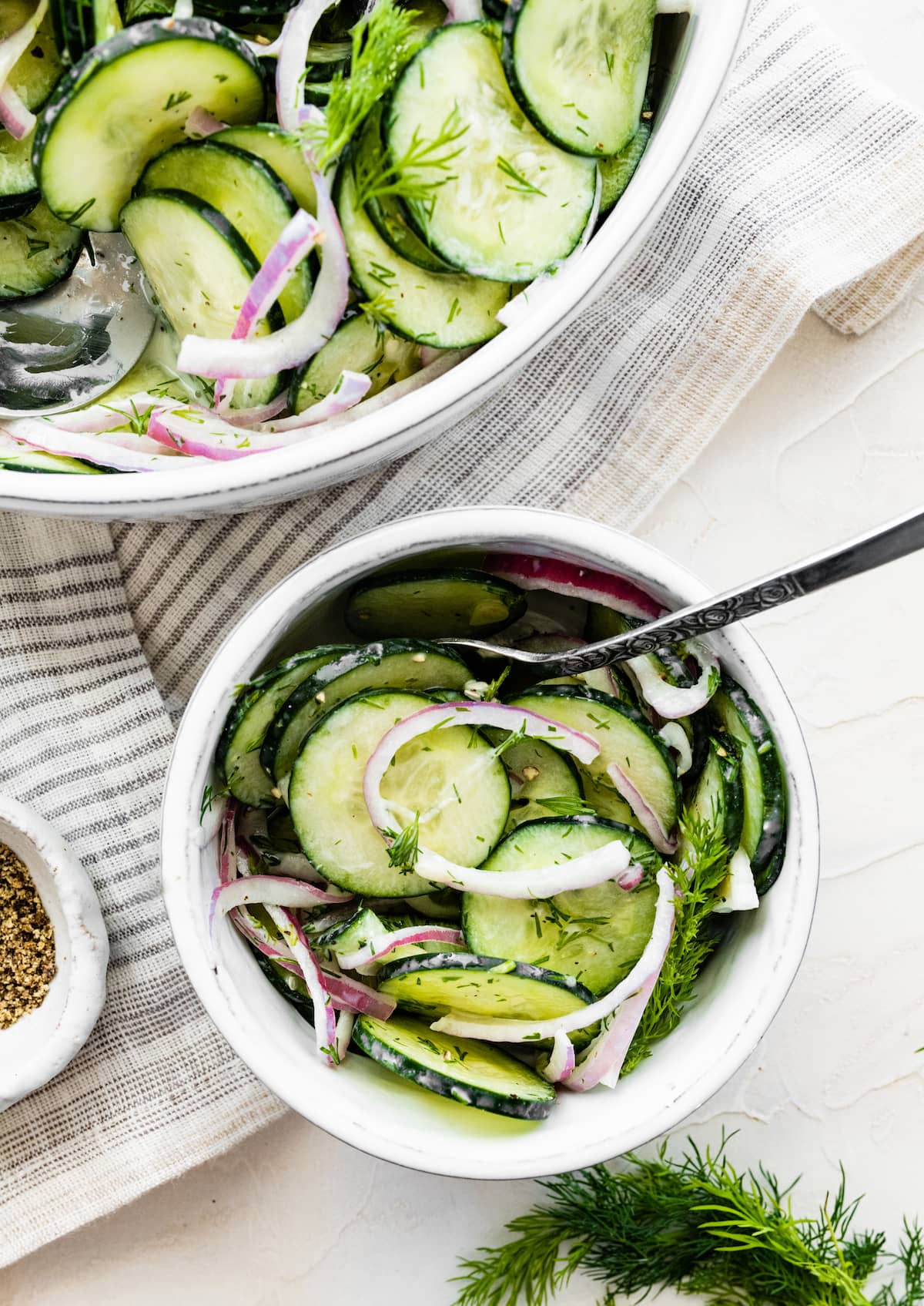 Creamy cucumber salad in a small white bowl with a metal fork near a larger bowl of the same salad.